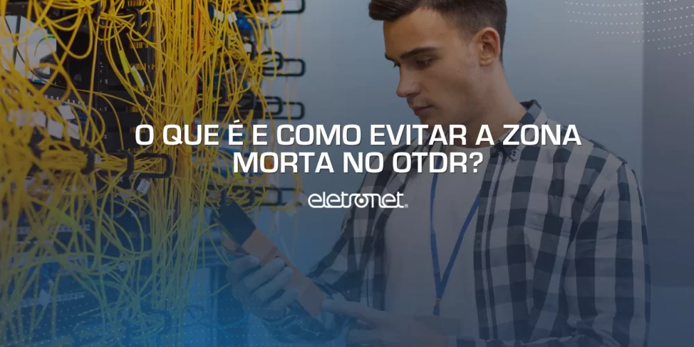 Homem branco com OTDR na mão medindo a zona morta da fibra óptica ao lado de um emaranhado de cabos amarelos.